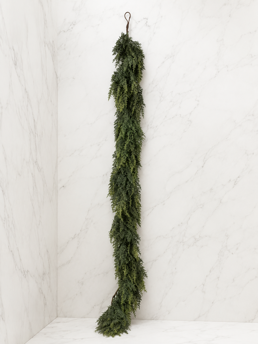 6-foot faux cedar garland displayed against a white marble studio backdrop, highlighting its realistic cascading greenery and natural texture.