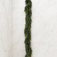 6-foot faux cedar garland displayed against a white marble studio backdrop, highlighting its realistic cascading greenery and natural texture.