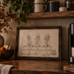 A framed sepia-toned art print featuring three cartoon skeletons sitting on a bench, styled on a rustic wooden countertop in a cozy kitchen setting with candles, plants, and a coffee machine.