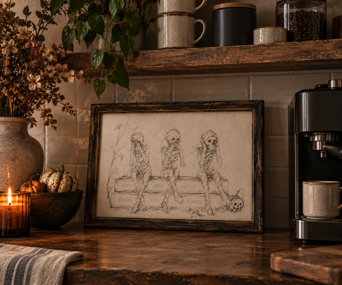 A framed sepia-toned art print featuring three cartoon skeletons sitting on a bench, styled on a rustic wooden countertop in a cozy kitchen setting with candles, plants, and a coffee machine.