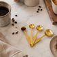 Gold stainless steel measuring spoon set styled on creamy ivory marble beside ceramic coffee cups, coffee beans, cocoa powder, and neutral stoneware in a warm California-modern editorial kitchen scene.