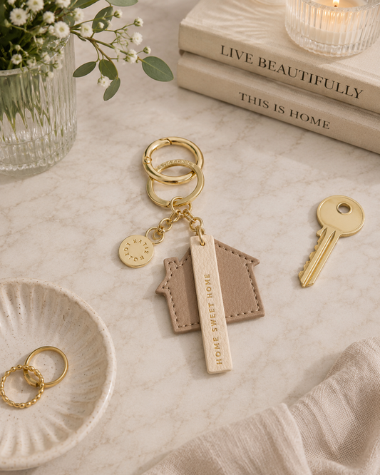 A gold-tone keychain set styled on a marble surface, featuring a house-shaped leather charm engraved with 'HOME SWEET HOME', a round wooden tag, a small circular charm, and a key beside it. Decorative props include books, a candle, flowers, and a dish with rings.