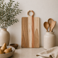 Oversized acacia wood serving board styled on a modern kitchen countertop with ceramic crock, wooden utensils, olive branches, and neutral décor.