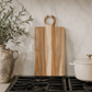 Oversized acacia wood serving board styled on a modern kitchen countertop with ceramic crock, wooden utensils, olive branches, and neutral décor.