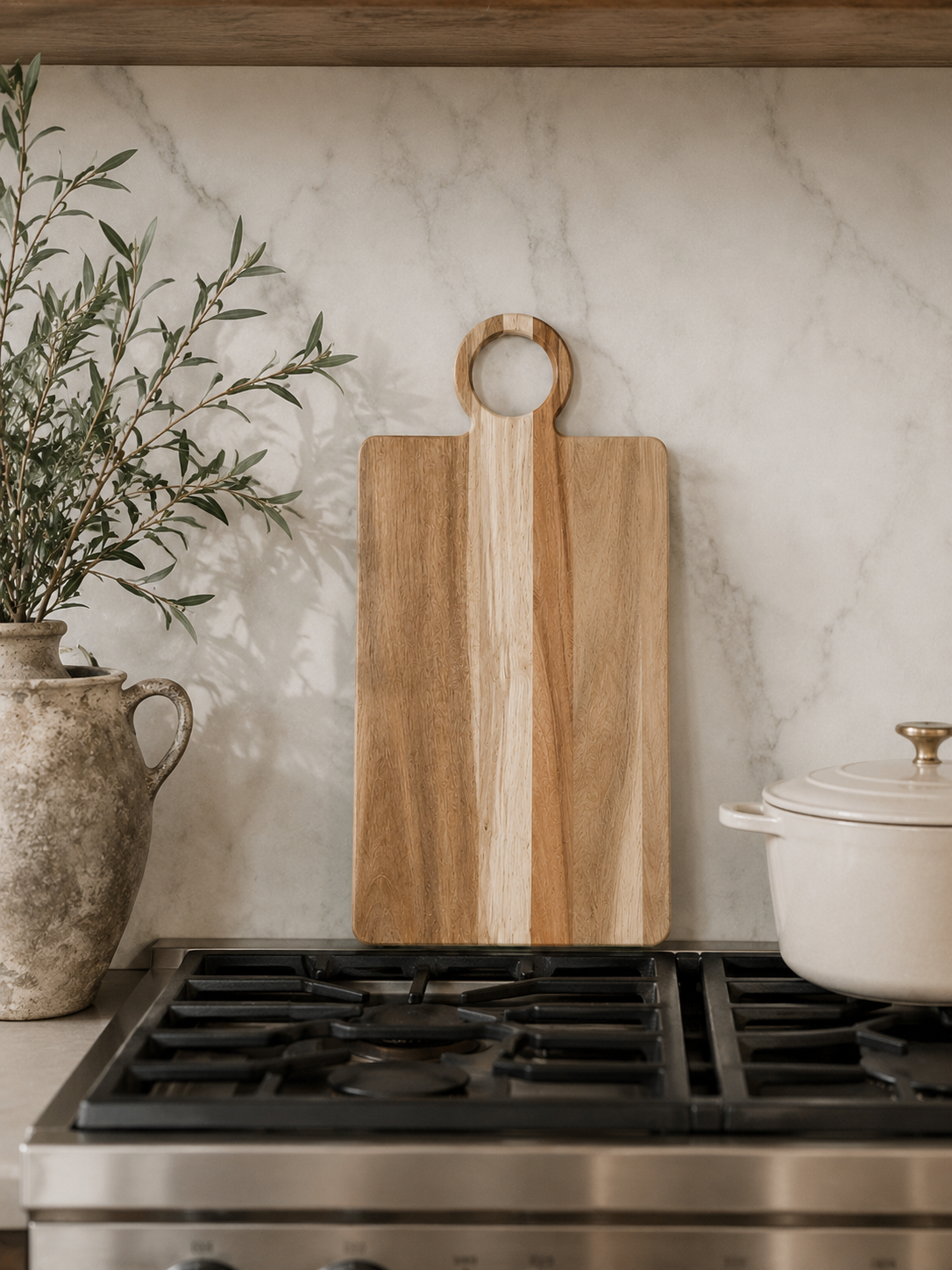 Oversized acacia wood serving board styled on a modern kitchen countertop with ceramic crock, wooden utensils, olive branches, and neutral décor.