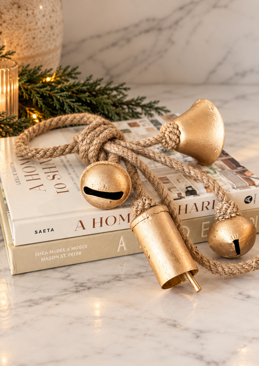 Antique gold Christmas bells featuring four assorted metal bells tied with natural jute rope, styled on a soft ivory marble background for timeless holiday decor.