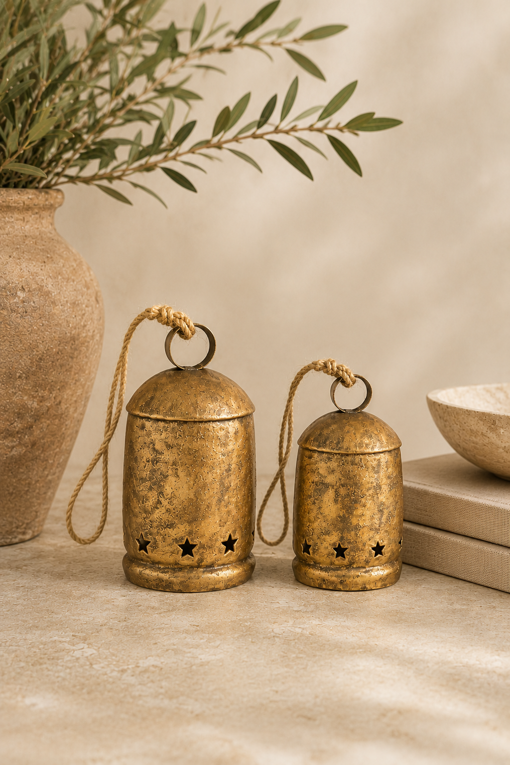 Large and medium antique gold hanging bells with distressed finish and star cutout details styled together in a warm designer holiday vignette with olive branches, neutral books, and natural stone accents.