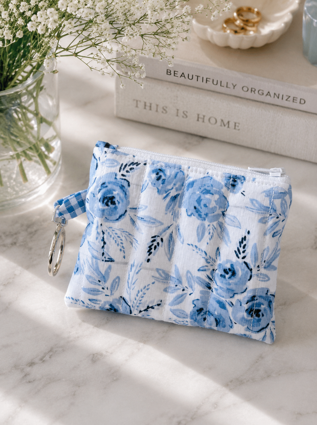 styled product photo of a blue floral quilted coin purse with silver key ring hardware on a marble countertop beside white flowers, neutral coffee table books, and soft luxury home decor accents in a bright coastal aesthetic.