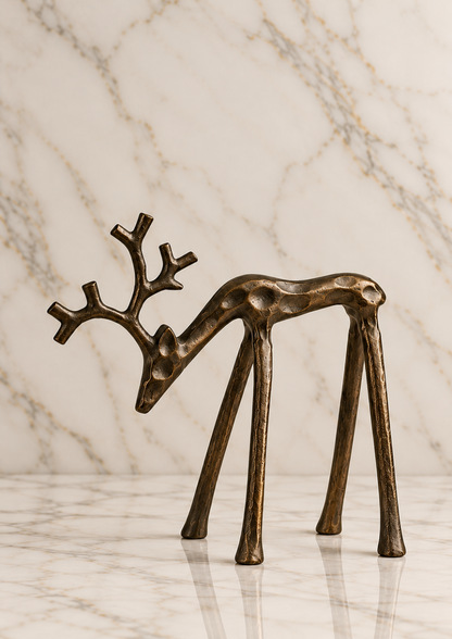 Grazing burnished iron reindeer sculpture featuring a gracefully lowered head, sculptural antlers, elongated legs, and an antiqued bronze-brown finish on an ivory marble studio background.