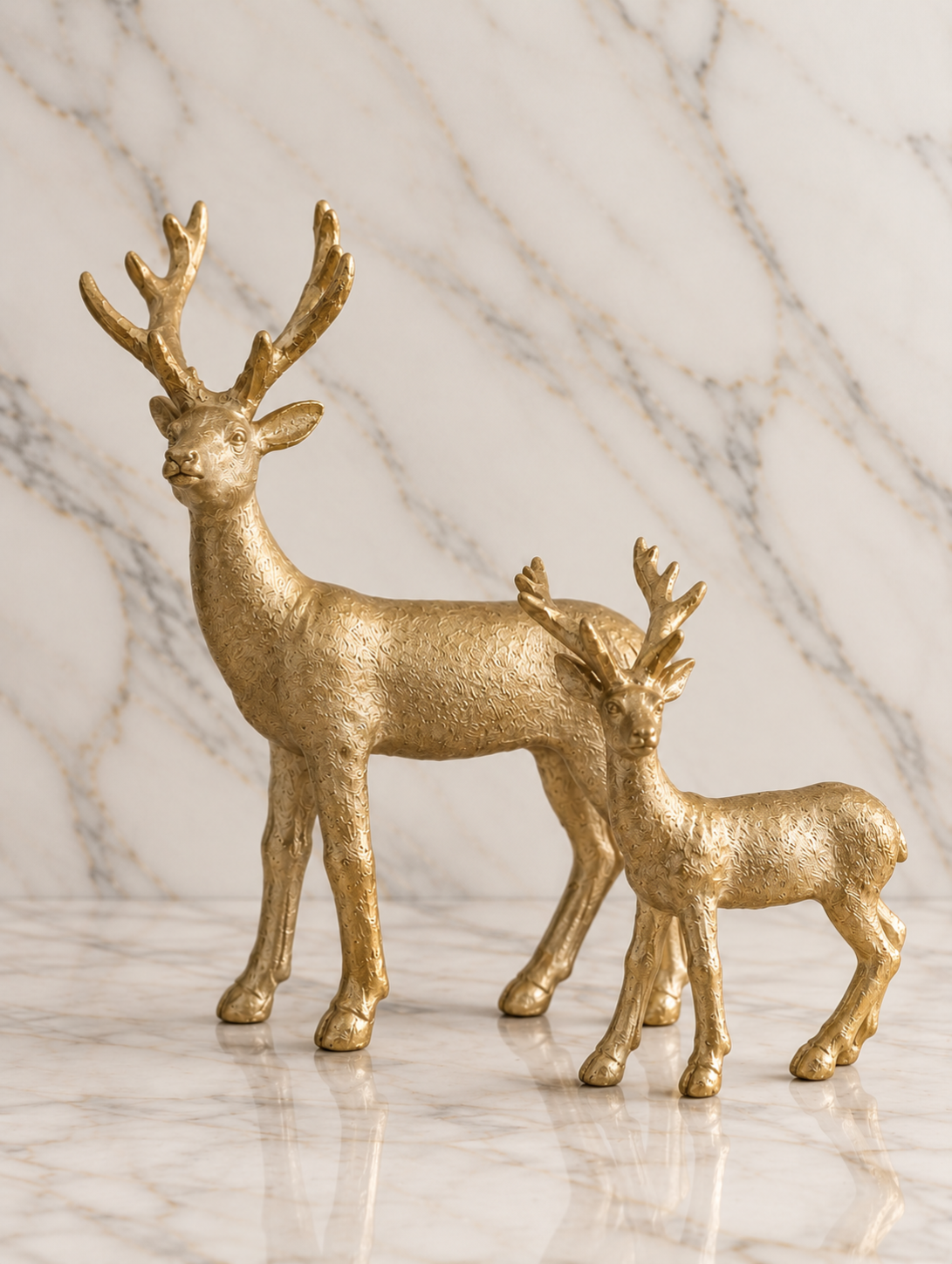 Set of two champagne gold reindeer sculptures in coordinating sizes, featuring textured metallic finishes and branching antlers on an ivory marble studio background.