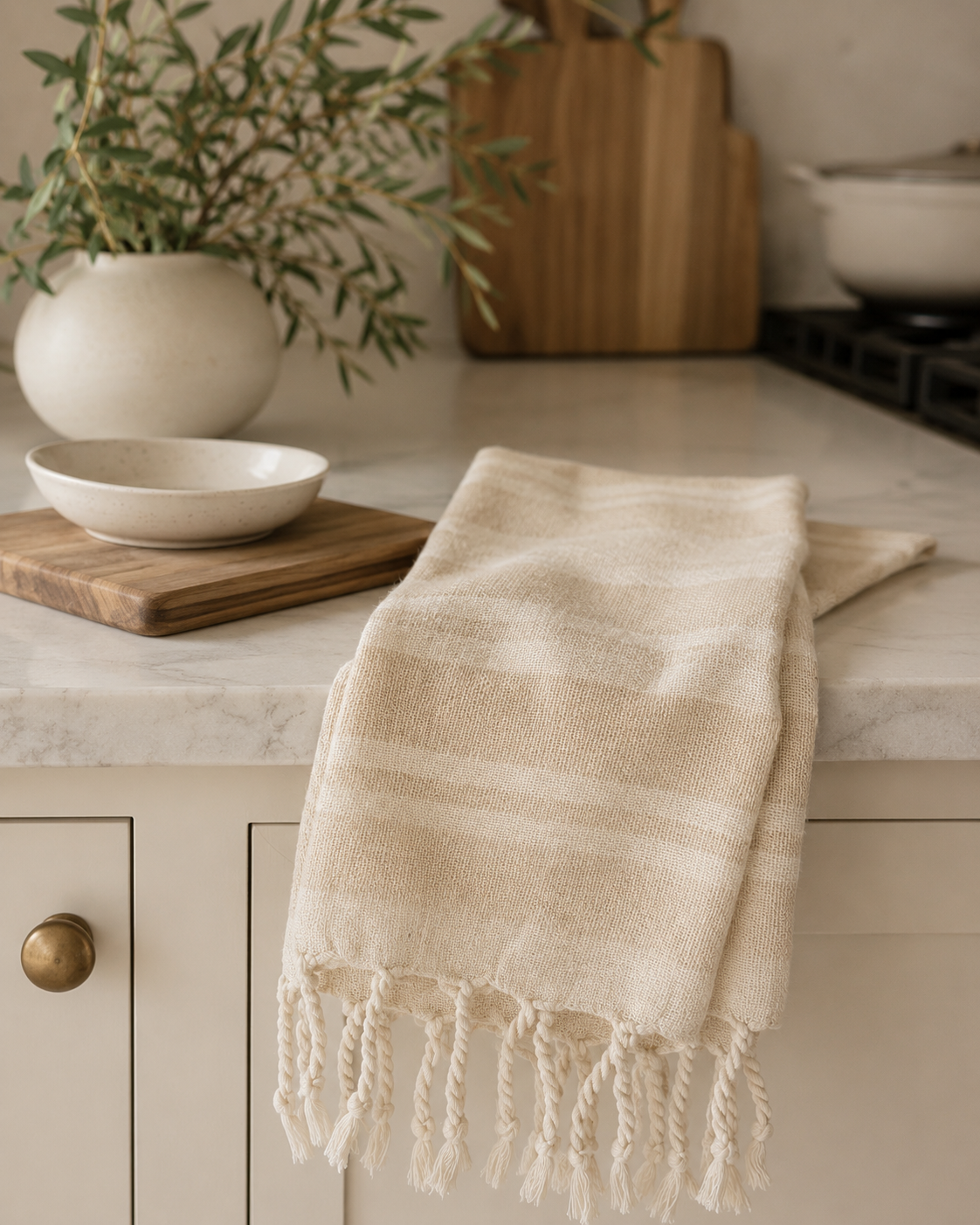 Cream striped woven Haman tea towel with decorative tassel fringe draped over a marble countertop in a modern California-inspired kitchen.