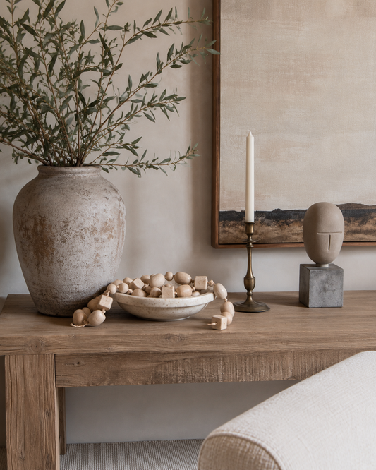 A styled interior scene with a large textured beige ceramic vase holding olive branches on a rustic wooden table, alongside a bowl of wooden beads, a brass candlestick with a white taper candle, and a sculptural wall piece.