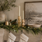 Creative Co-Op mango wood candlesticks with ivory taper candles styled on a Christmas mantel with fresh cedar and eucalyptus garland, warm white fairy lights, vintage brass bells, a neutral winter landscape, stoneware vase, marble bowl, and knit stockings in a designer-inspired holiday setting.
