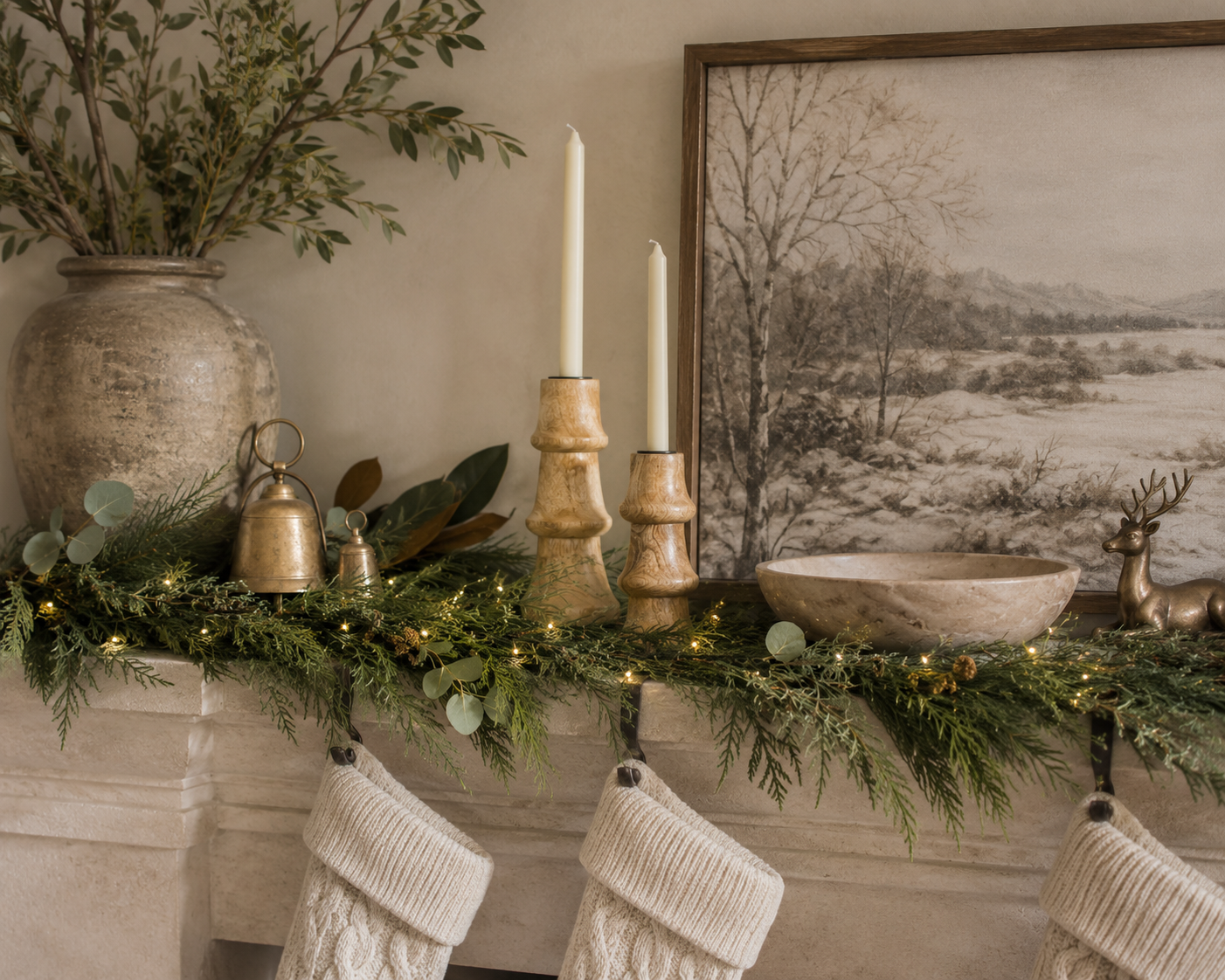 Creative Co-Op mango wood candlesticks with ivory taper candles styled on a Christmas mantel with fresh cedar and eucalyptus garland, warm white fairy lights, vintage brass bells, a neutral winter landscape, stoneware vase, marble bowl, and knit stockings in a designer-inspired holiday setting.