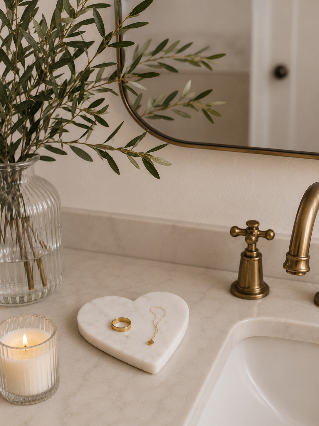 White marble heart-shaped trinket dish styled on a bathroom vanity with jewelry, a glass vase, and greenery.