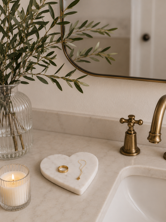 White marble heart-shaped trinket dish styled on a bathroom vanity with jewelry, a glass vase, and greenery.