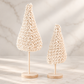 rochet Christmas trees on natural wood stands styled against a soft neutral marble backdrop.