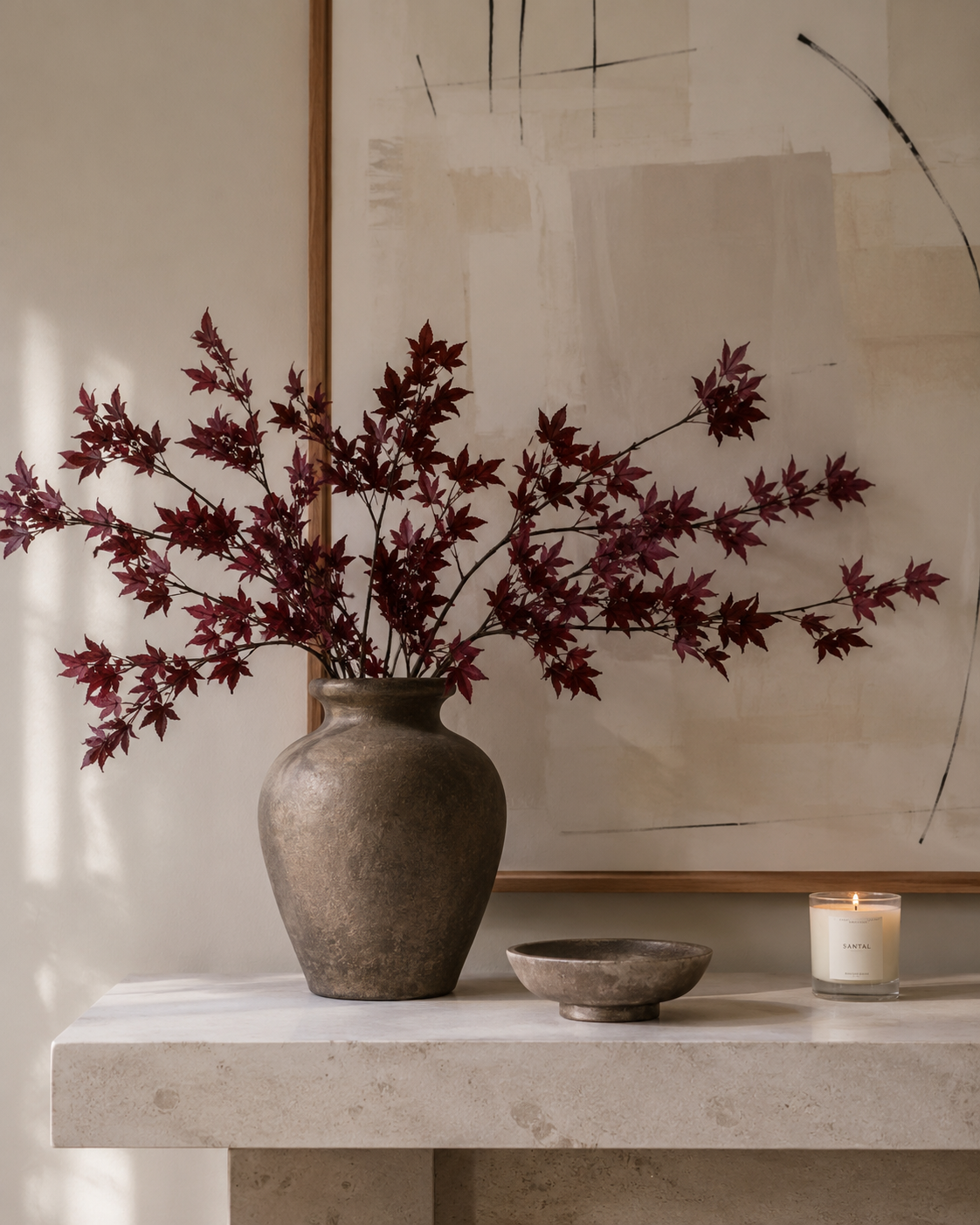 Dark red faux maple leaf stems arranged in a ceramic vase on a stone console light painting and modern organic décor.