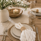 White fringed linen napkin with a woven napkin ring styled on a neutral tablescape with ceramic dinnerware, woven placemats, candles, and greenery in a modern coastal dining room.
