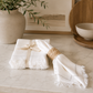 Set of white fringed linen napkins with decorative stitched edging styled on a marble countertop with a woven napkin ring in a modern California-inspired kitchen.