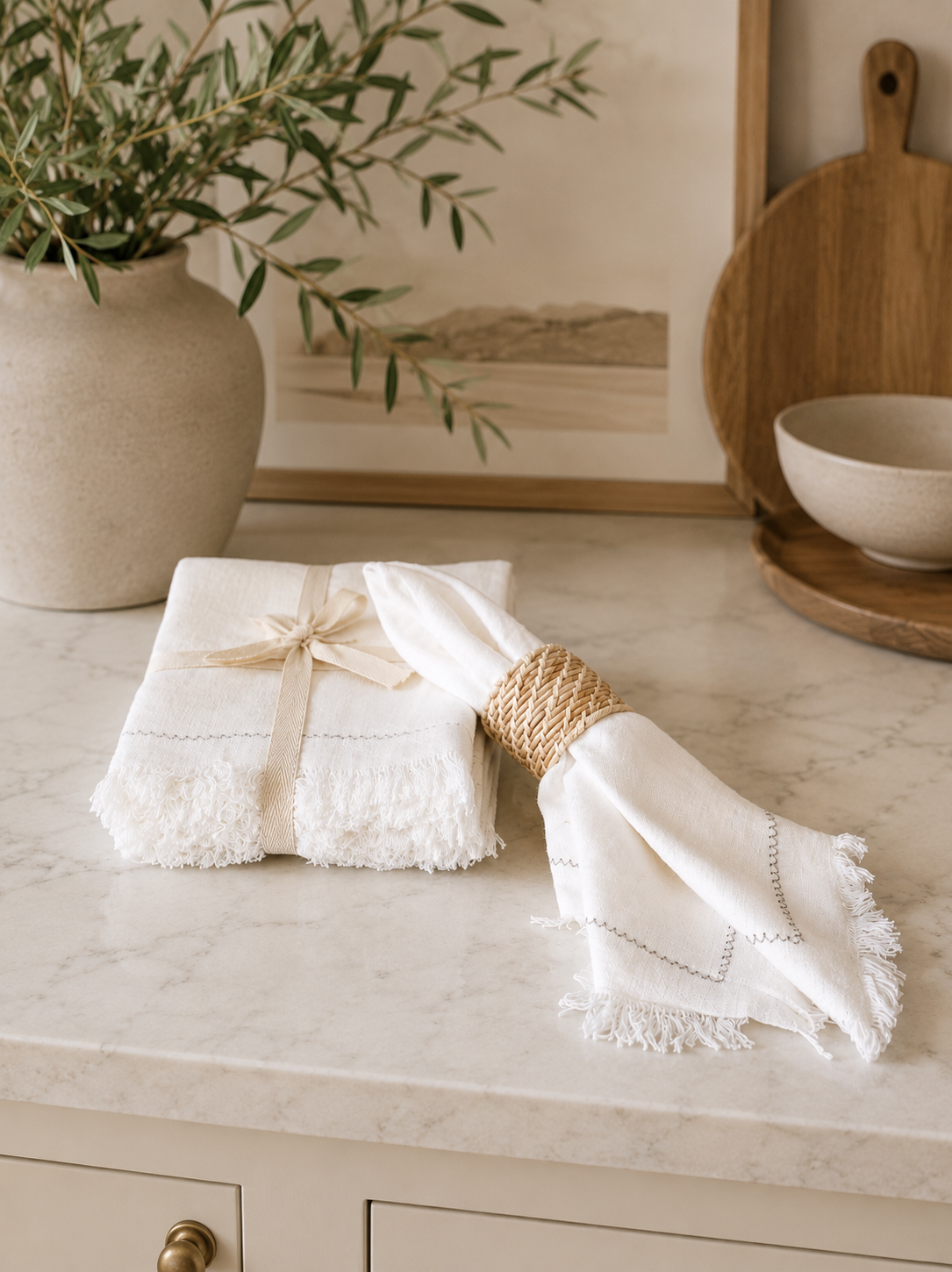 Set of white fringed linen napkins with decorative stitched edging styled on a marble countertop with a woven napkin ring in a modern California-inspired kitchen.