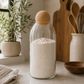Tall glass storage jar with cork ball lid filled with flour, styled on a modern kitchen countertop with ceramic utensils, wooden cutting boards, and natural greenery.