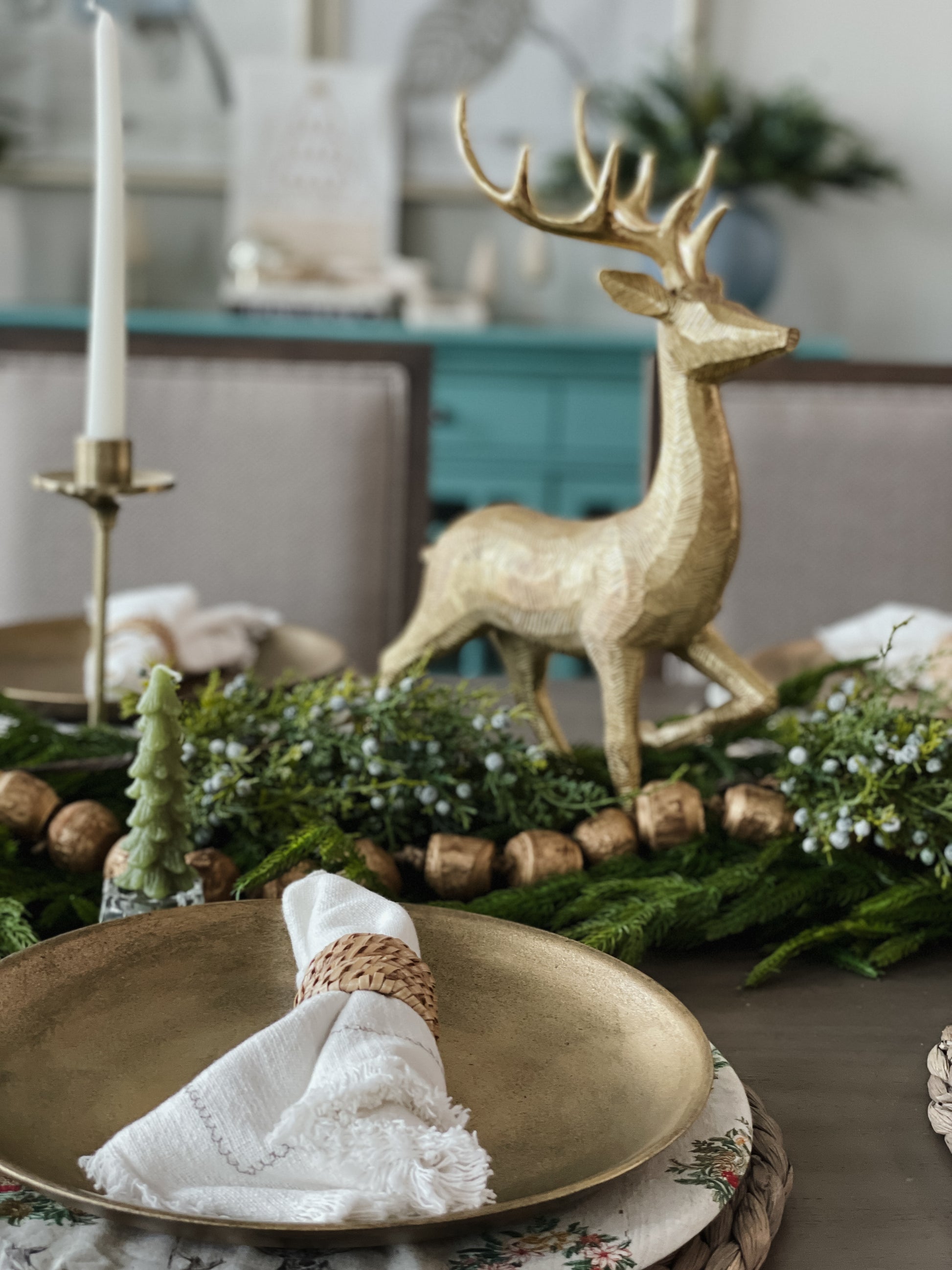 Gold finish decorative plate styled on a holiday tablescape with a white napkin, greenery garland and a gold reindeer centerpiece