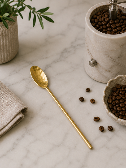 Gold hammered stainless steel coffee spoon styled beside a marble coffee grinder with whole coffee beans on a white marble countertop in a modern California-inspired kitchen.