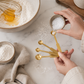 Gold stainless steel measuring spoon set being used in a warm California-modern baking scene with sea salt, ceramic mixing bowls, flour, linen textiles, and creamy ivory marble countertop.