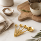 Gold stainless steel measuring spoon set styled on creamy ivory marble beside neutral ceramic bowls, folded linen, olive branches, sea salt, and warm oak cutting board in a California-modern editorial kitchen scene.