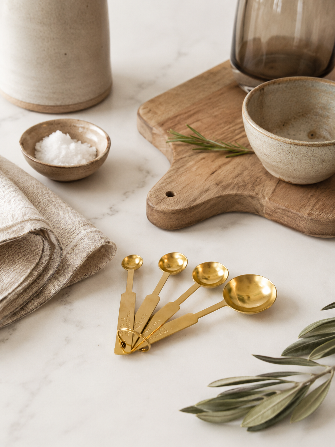 Gold stainless steel measuring spoon set styled on creamy ivory marble beside neutral ceramic bowls, folded linen, olive branches, sea salt, and warm oak cutting board in a California-modern editorial kitchen scene.