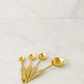 olished gold stainless steel measuring spoon set arranged in a minimal fanned composition on creamy ivory marble with soft diffused studio lighting.