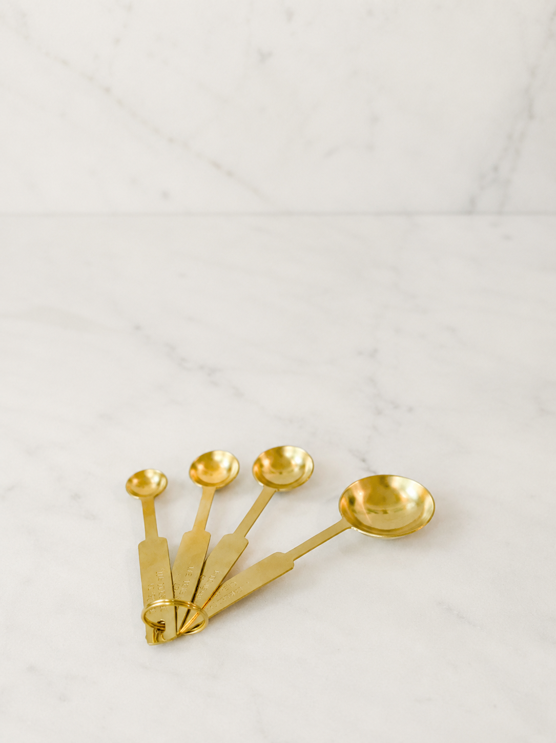 olished gold stainless steel measuring spoon set arranged in a minimal fanned composition on creamy ivory marble with soft diffused studio lighting.