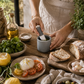 Petite gray ceramic mortar and pestle being used to prepare fresh basil pesto on a rustic wood table surrounded by lemons, burrata, olives, artisan bread, and layered California coastal hosting decor.
