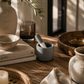 Small gray ceramic mortar and pestle styled on a rustic wood table with neutral pottery, linen textiles, olive branches, and warm California-modern decor