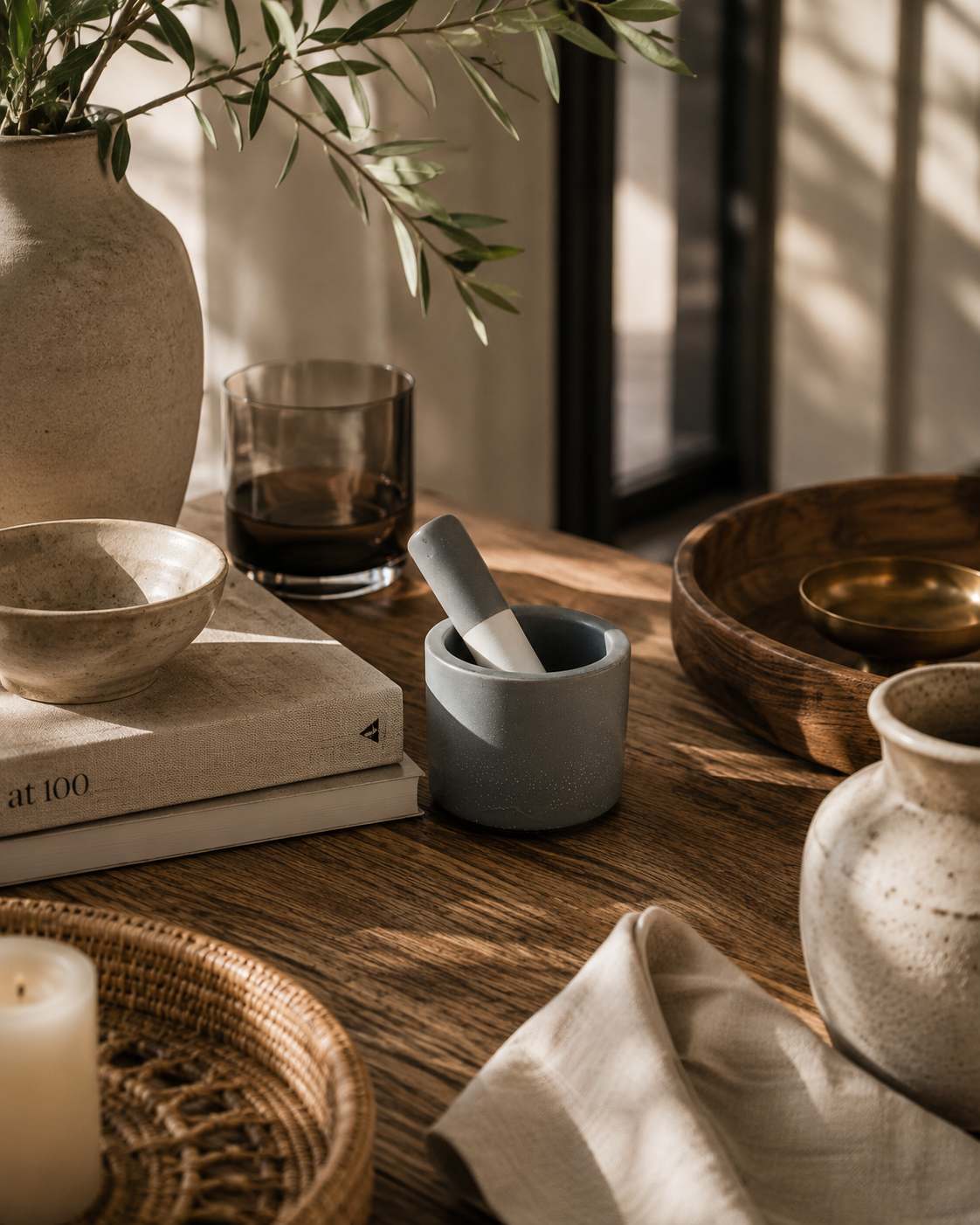 Small gray ceramic mortar and pestle styled on a rustic wood table with neutral pottery, linen textiles, olive branches, and warm California-modern decor