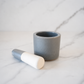 Small gray speckled ceramic mortar and pestle with white-tipped pestle on a white marble background in a clean neutral studio setting.