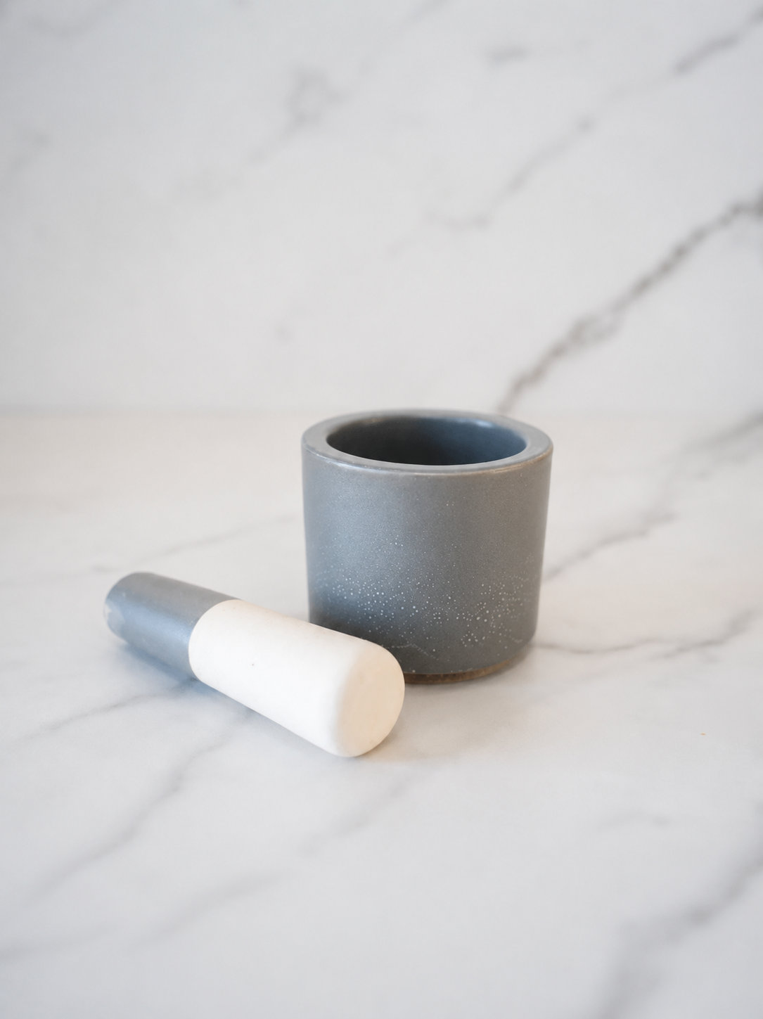 Small gray speckled ceramic mortar and pestle with white-tipped pestle on a white marble background in a clean neutral studio setting.