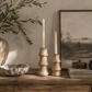 Two textured mango wood candlestick holders with white taper candles, styled on a wooden console table next to a large vase, a stone bowl, a framed landscape print, and books.