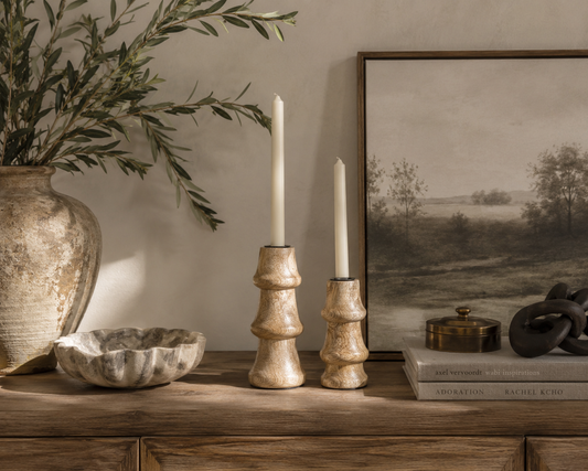 Two textured mango wood candlestick holders with white taper candles, styled on a wooden console table next to a large vase, a stone bowl, a framed landscape print, and books.