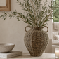 Decorative woven vase with green branches on a table in a room setting.