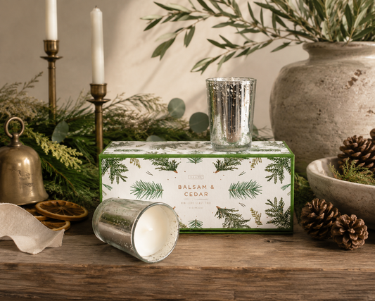 Illume Balsam & Cedar mercury glass votive candle gift set featuring three 2.5-inch scented candles in silver mercury glass holders, beautifully styled with fresh cedar greenery, pinecones, brass accents, and natural holiday décor.