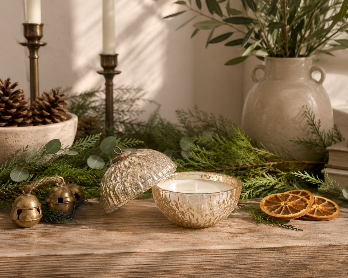 illume winter white mercury glass ornament candle with removable lid styled on a reclaimed wood table with fresh cedar and eucalyptus greenery, vintage brass bells, dried orange slices, pinecones, and warm neutral holiday décor in a designer-inspired Christmas setting.