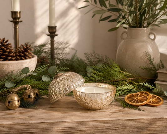 illume winter white mercury glass ornament candle with removable lid styled on a reclaimed wood table with fresh cedar and eucalyptus greenery, vintage brass bells, dried orange slices, pinecones, and warm neutral holiday décor in a designer-inspired Christmas setting.