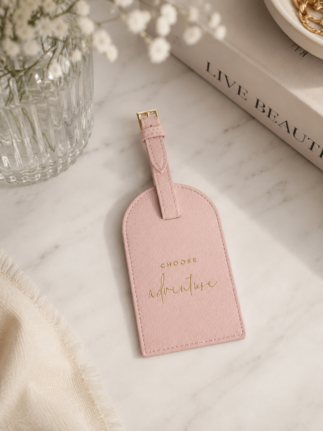 Katie Loxton Choose Adventure luggage tag in Dusty Pink faux leather styled on a neutral marble surface with elegant travel-inspired décor.