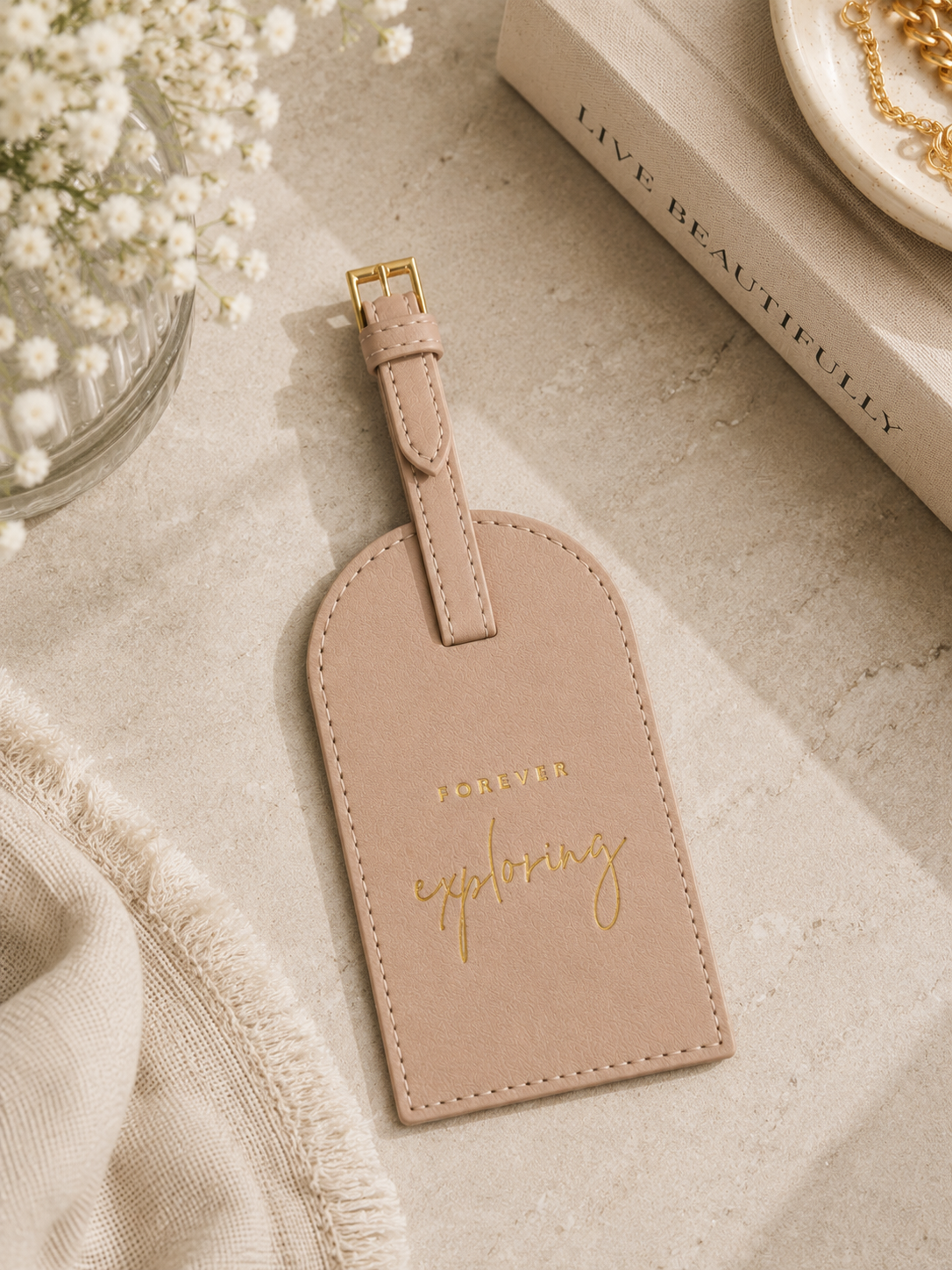 Katie Loxton Forever Exploring luggage tag in Soft Tan faux leather styled on a neutral tabletop with elegant travel accessories and soft natural lighting.