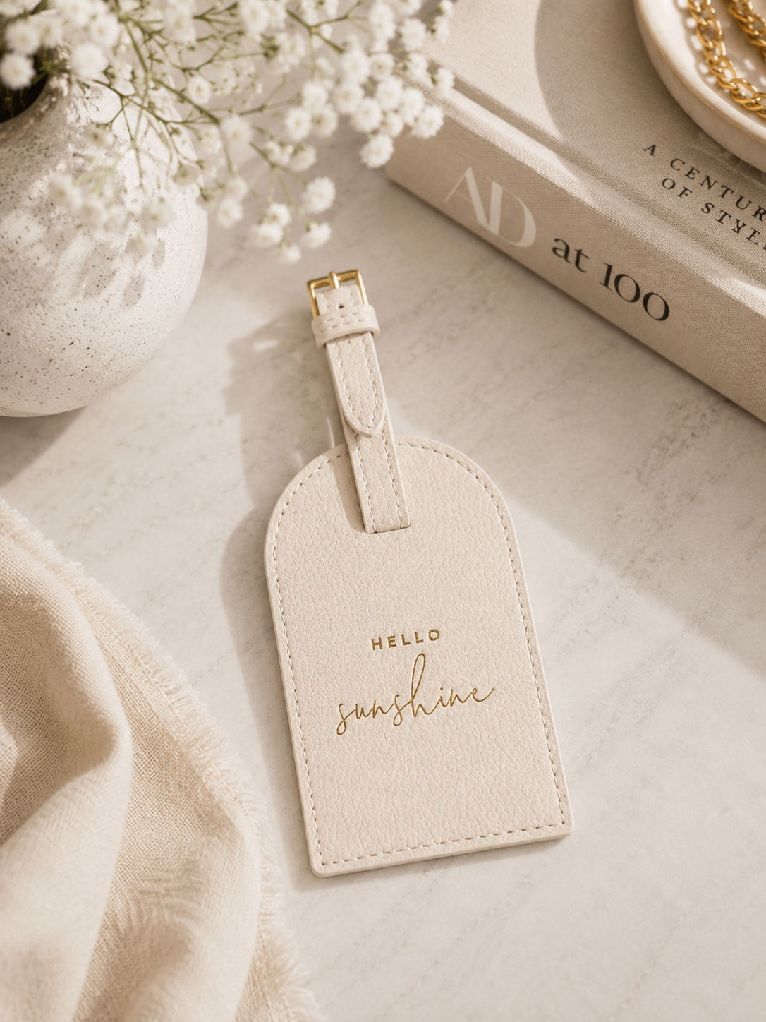 Katie Loxton Hello Sunshine luggage tag in Eggshell faux leather styled on a neutral marble surface with elegant travel-inspired décor.