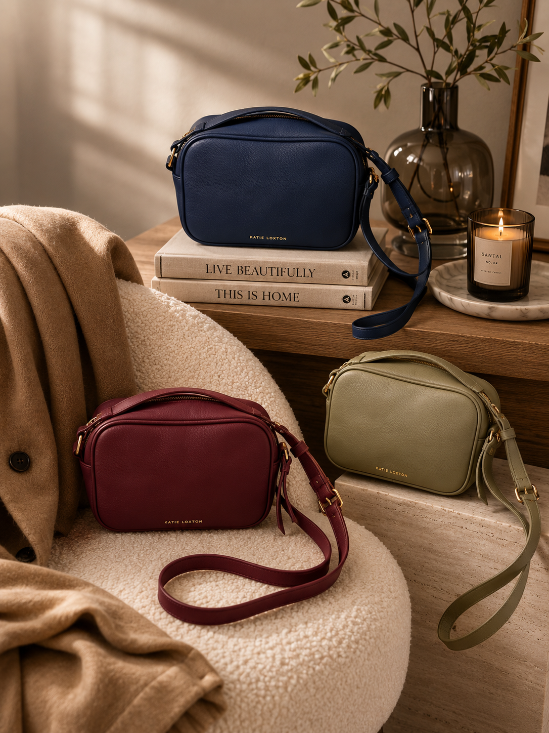 Katie Loxton Isla crossbody handbags in burgundy, navy, and sage green styled in a warm neutral living room with luxury editorial decor, soft lighting, and modern organic interiors.