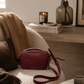 Katie Loxton Isla burgundy crossbody bag styled in a warm neutral luxury interior with soft editorial lighting, oak textures, and elevated California-modern decor.