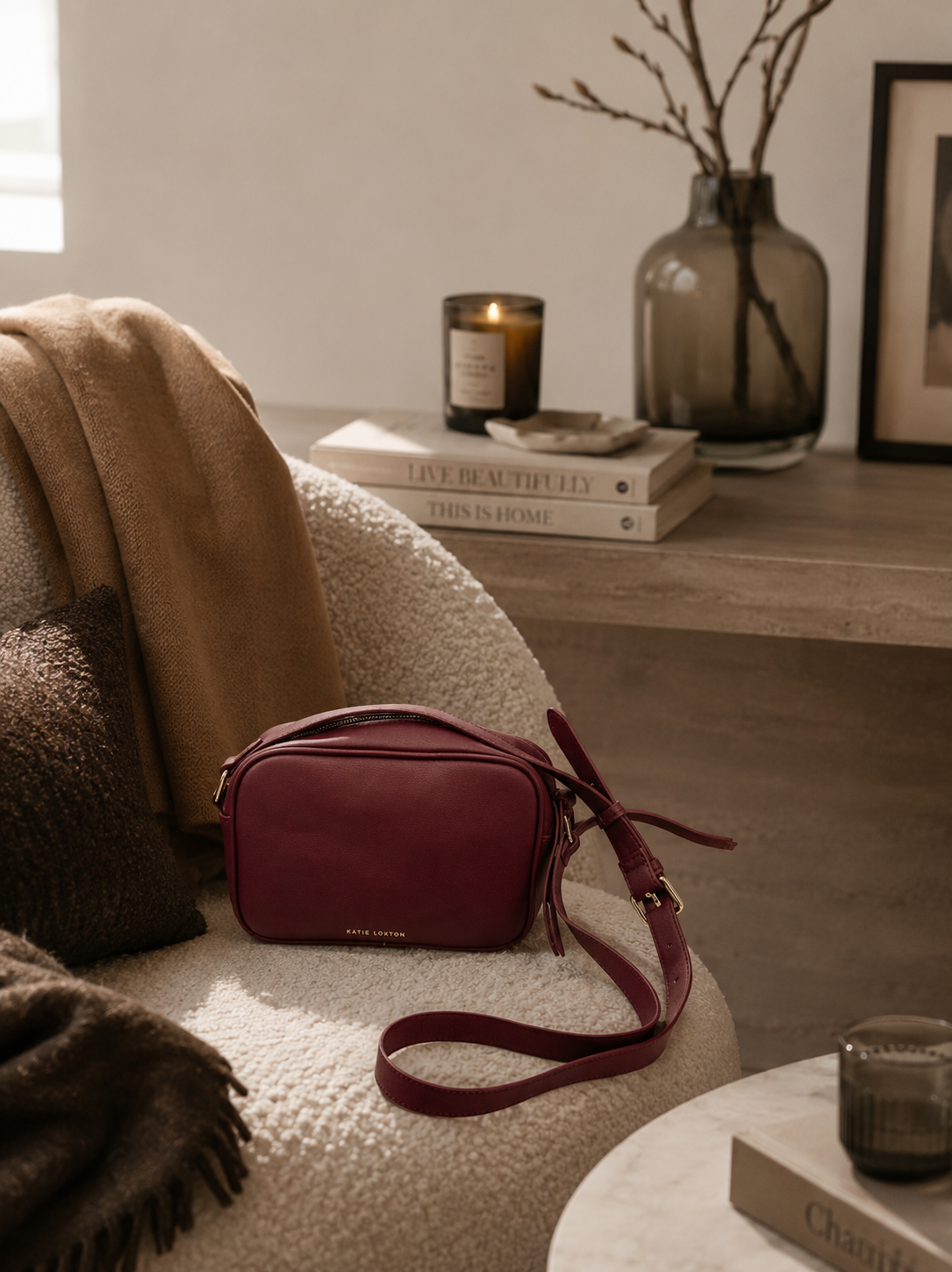 Katie Loxton Isla burgundy crossbody bag styled in a warm neutral luxury interior with soft editorial lighting, oak textures, and elevated California-modern decor.